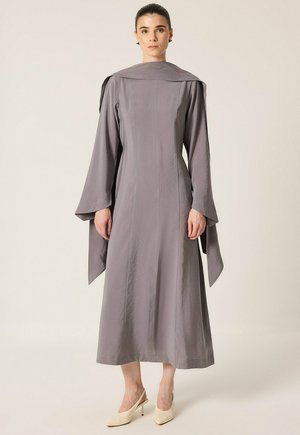 Femme portant une longue robe grise à larges manches avec un détail assorti aux épaules, associée à des escarpins beiges à bouts pointus, debout devant un fond uni.