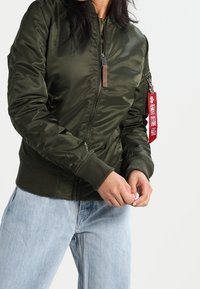 Oliwkowa bomberka z gładkim, błyszczącym wykończeniem, ściągaczami na mankietach i z zamkiem błyskawicznym z przodu. Posiada markową czerwoną metkę na zamku.