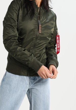 Veste bomber vert olive avec une finition lisse et brillante, des poignets côtelés et une fermeture éclair à l'avant. Comprend une étiquette de marque rouge sur la fermeture éclair.
