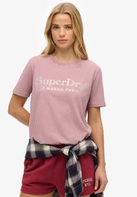 Lyserød bomulds T-shirt med korte ærmer, prydet med "SuperDry" logo i metallisk sølv. Båret med mørkerøde shorts og en ternet skjorte bundet om livet.
