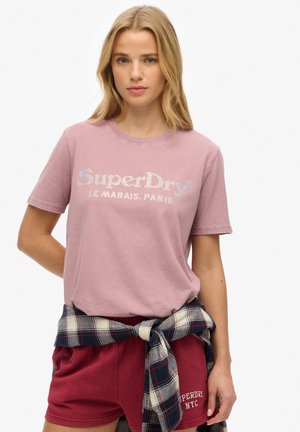 Camiseta de algodón rosa claro con mangas cortas que presenta el logo "SuperDry" en plateado metálico. Llevada con pantalones cortos burdeos y una camisa de cuadros atada en la cintura.
