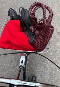 Sandali neri, un maglione rosso lavorato a maglia e una borsa intrecciata bordeaux nel cestino di una bicicletta. Manubrio metallico parzialmente visibile sotto.
