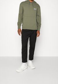 Uomo che indossa un felpa verde oliva, pantaloni neri, calze bianche e sneaker bianche, in piedi contro uno sfondo bianco uniforme.