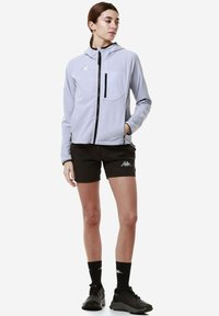 Giacca a cappuccio grigio chiaro con tasca a zip sul petto, zip nera e motivo a rete, abbinata a pantaloni corti neri e calze e scarpe nere.