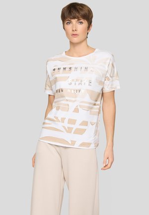 Frau mit kurzen braunen Haaren, die ein weißes und beiges gemustertes T-Shirt mit Text und beige weite Hosen trägt, steht vor einem grauen Hintergrund.