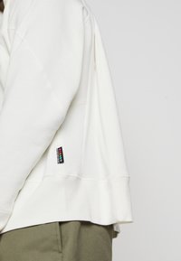 Bílá mikina s kapucí vyrobena z měkké látky, s kratším střihem, bočními rozparky a barevnou značkovou visačkou na levé straně.