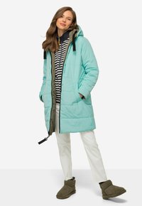 Giacca lunga in piuma turchese con foderato grigio, dettagli neri e tasche. Indossata sopra un hoodie a righe blu e bianche e pantaloni bianchi.