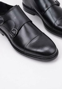 Zapatos de cuero negro con puntera redonda, diseño de doble hebilla y acabado suave. Presentan un tacón bajo y detalles de costura en contraste.