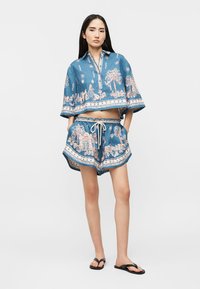 Frau trägt ein blau gemustertes Croptop und passende Shorts mit Kordelzug, steht mit den Händen in den Taschen und trägt schwarze Flip-Flops.