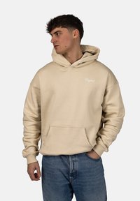 Beige mikina s kapucí vyrobená z měkkého materiálu, se přední kapsou a žebrovanými manžetami. Umístěné logo v bílé barvě, volný střih.