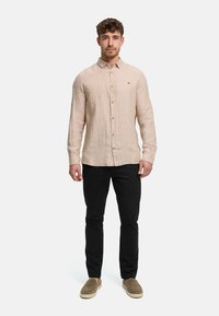 Man standing facing forward wearing a beige button-up linen shirt, black pants, and brown slip-on shoes on plain background.