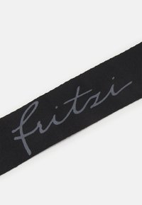 Czarny materiałowy pasek z napisem "fritzi" w szarym, kursywnym kroju pisma, prezentujący miękką teksturę i prosty, niewyszukany design.