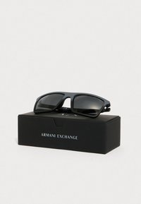 Ochelari de soare negri cu un finisaj mat, lentile dreptunghiulare și o marcă subtilă, așezați pe o cutie neagră etichetată "Armani Exchange".
