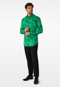 Mann richtet die Manschette eines langärmligen, grünen Hemdes mit abstraktem Muster, trägt schwarze Hose und schwarze Lederschuhe, steht vor weißem Hintergrund.