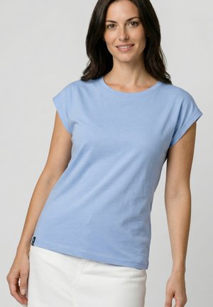 Frau mit schulterlangem dunklem Haar, die ein hellblaues kurzärmliges Oberteil und einen weißen Rock trägt, leicht lächelnd, vor einem schlichten Hintergrund.