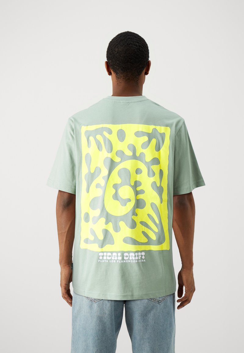 Camiseta de manga corta en verde menta con un gran gráfico abstracto amarillo en la parte posterior y texto blanco en la parte inferior, hecha de material de algodón.