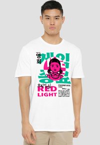 A young man stands wearing a white t-shirt featuring colorful text and an illustration of a girl, referencing a game context.