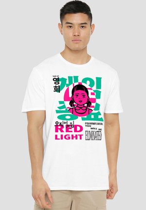 Ein junger Mann steht in einem weißen T-Shirt, das bunten Text und eine Illustration eines Mädchens zeigt, die auf einen Spielkontext anspielt.