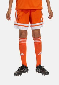 Orange Sportshorts mit weißen Streifen, kombiniert mit orangefarbenen kniehohen Socken mit weißen Akzenten. Schwarze Fußballschuhe mit strukturiertem Design.