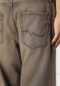 Pantalon en denim marron avec une surface texturée, comprenant deux poches arrière et des détails de couture contrastante le long des bords des poches.