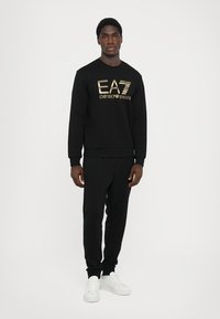 Svart bomull sweatshirt med gull "EA7 Emporio Armani" logo, parret med svarte bukser og hvite sneakers. Enkelt atletisk design.