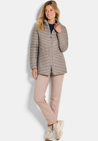 Vrouw in een taupe gewatteerde jas, lichtroze broek en beige sneakers, staand met één been gekruist en met een subtiele glimlach naar de zijkant.