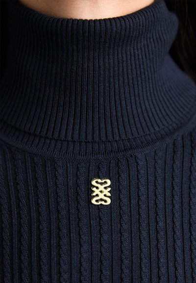 Marineblå ribbet turtleneck sweater i strikstof med et subtilt tekstureret mønster og en gylden dekorativ detalje.