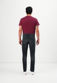 Mann in dunkelgrauen Jeans, einem bordeauxfarbenen T-Shirt und weißen Sneakers, der mit dem Rücken zu einer weißen Wand steht.