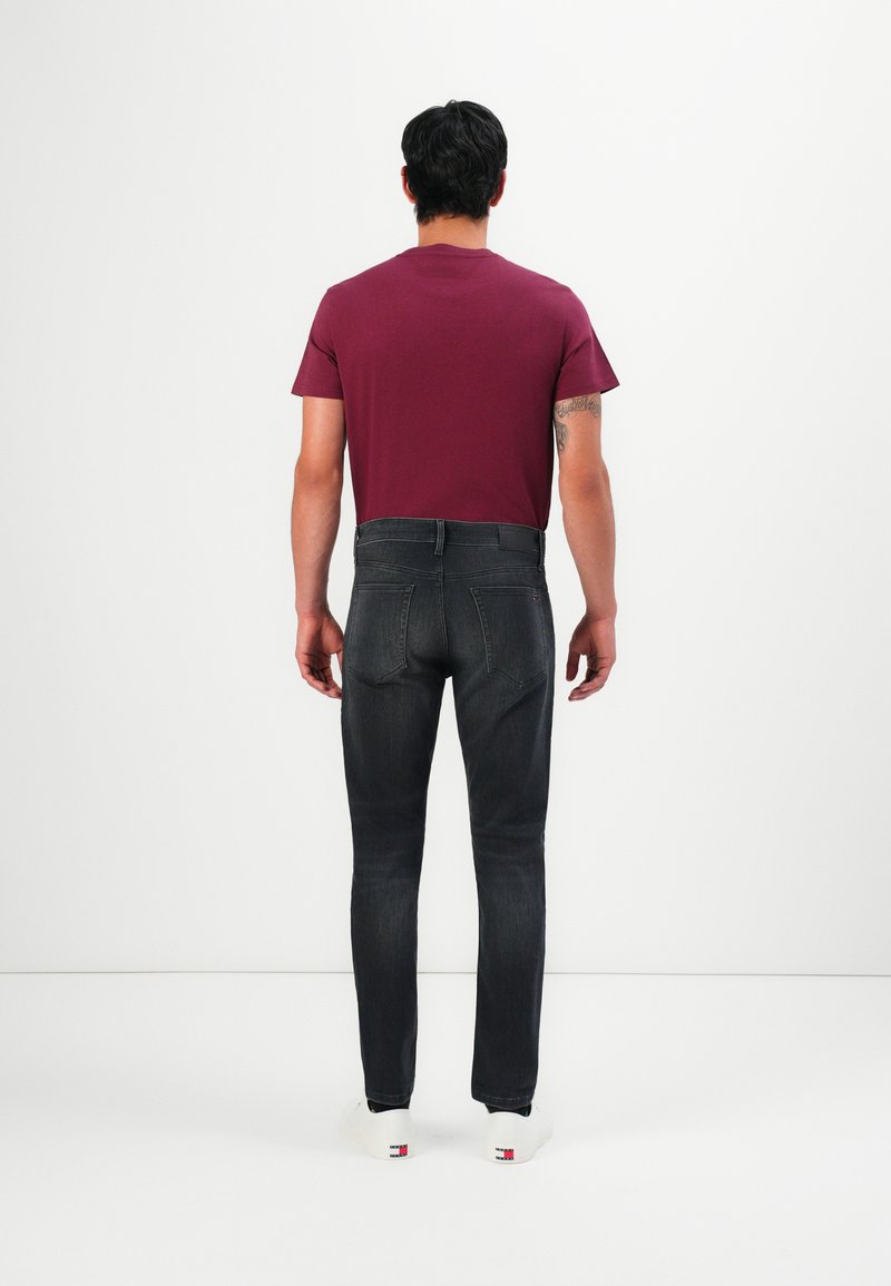 Man wearing dark gray jeans, a maroon t-shirt, and white sneakers standing facing a white wall, showing back view.