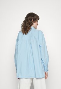 Camisa oversized azul clara com colarinho abotoado, mangas longas e uma vincha nas costas para adicionar volume e movimento.