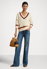 Kremasta pulover s uzorkom pletenja u obliku kabela s narančastim i tamnoplavim prugama na V-izrezu i rubu, uparena s plavim trapericama širokih nogavica i smeđom torbom.