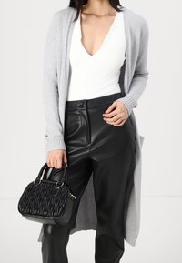 Grauer langer Cardigan über einem weißen Top, kombiniert mit schwarzen Kunstlederhosen. Eine schwarze gesteppte Handtasche mit runden Griffen wird in einer Hand gehalten.