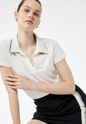 Valkoinen polo-paita mustilla pipeillä, lyhyet hihat ja V-kaula-aukko. Yhdistetty mustan hameen kanssa, jossa on sivuraidat ja muotoiltu design.