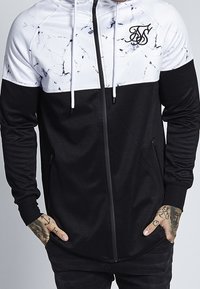 Svart och vit jacka med ett marmormönster på toppen, med dragkedja fram, två sidofickor och en logotyp på bröstet.