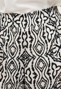 Ύφασμα με γεωμετρικό σχέδιο σε μαύρο και μπεζ χρώμα σε πλισέ παντελόνια φαρδιάς γραμμής, φορεμένα με λευκό τοπ μέσα στη μέση.