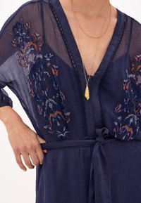 Robe de style kimono bleu marine avec broderies florales en orange et bleu. Présente un décolleté en V profond, un tissu transparent et un détail de taille nouée.