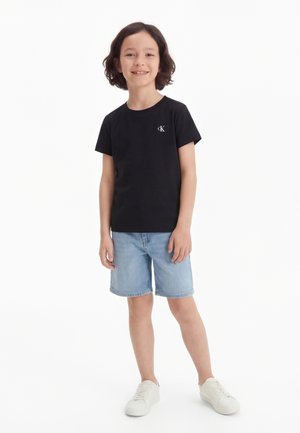 Copil zâmbitor în picioare, purtând un tricou negru Calvin Klein, pantaloni scurți din denim albastru deschis și adidași albi pe un fundal alb.