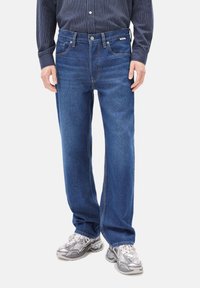 Jeans de denim azul con corte recto, diseño de cinco bolsillos y detalles de costura visibles. Llevados con una camisa a rayas y zapatillas.