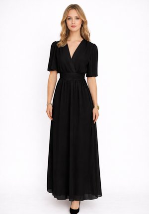 Mujer con vestido negro hasta el suelo, de manga corta y escote en V, de pie frente a un fondo blanco.