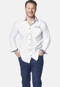 Weißes Hemd mit schwarzem Knopf, kontrastierenden Manschetten und Taschen Details. Kombiniert mit dunklen Jeans. Modell steht mit Händen in den Taschen.