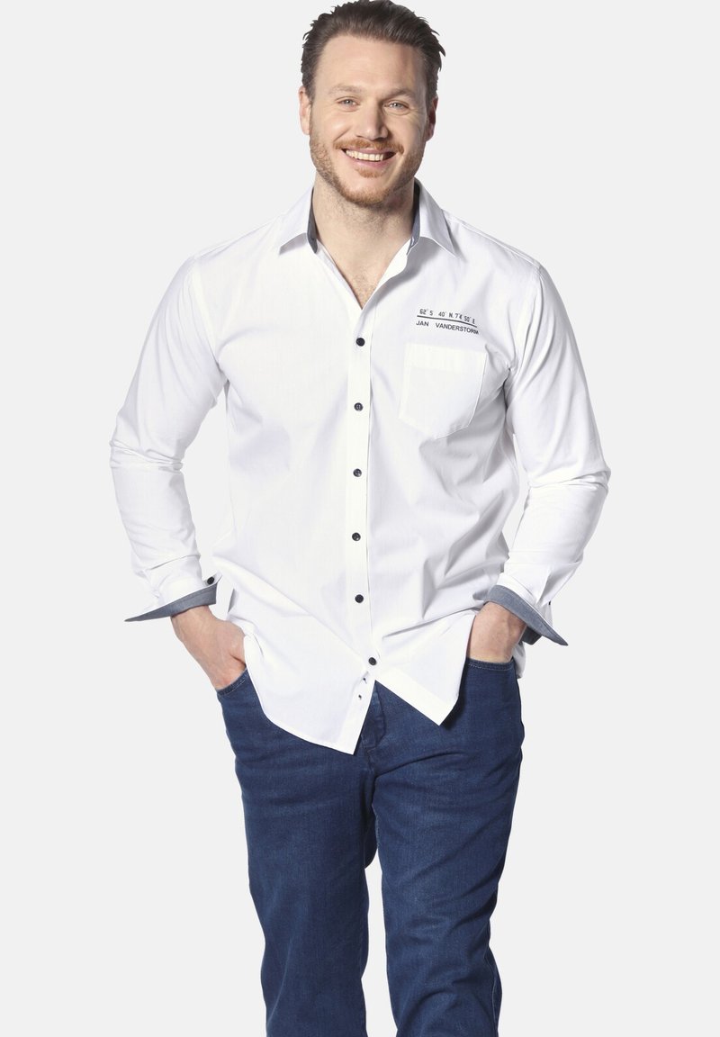 Weißes Hemd mit schwarzem Knopf, kontrastierenden Manschetten und Taschen Details. Kombiniert mit dunklen Jeans. Modell steht mit Händen in den Taschen.