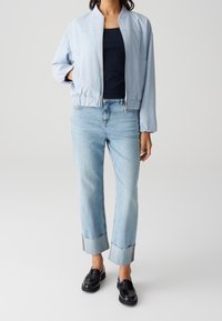 Femme portant une veste zippée bleu clair, un haut bleu marine, un jean clair retroussé et des mocassins noirs, debout devant un fond uni.