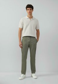 Helles graues, strukturiertes Poloshirt mit Kragen; olivgrüne Hose; weiße Sneakers; stehende Pose vor einem neutralen Hintergrund.