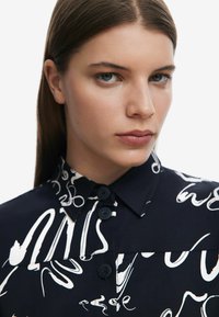 Maglietta navy con motivo astratto bianco, caratterizzata da un colletto classico, tre bottoni e una texture liscia. Primo piano del viso del modello.