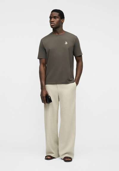 Άνδρας που φοράει ελιάς κοντομάνικο πουκάμισο με μικρό λογότυπο ζώου, μπεζ παντελόνι φαρδύ, μαύρα σανδάλια, κρατώντας ένα μαύρο πορτοφόλι.