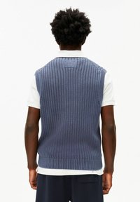 Mouwloze gebreide pullover in marineblauw met geribbelde structuur; heeft een ronde halslijn en wordt gedragen over een wit overhemd met kraag.