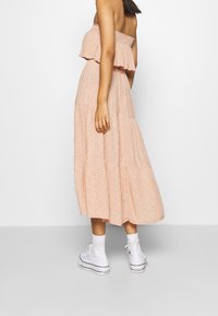 Robe maxi rose à motifs, dénudée des épaules, avec des volants, portée avec des baskets montantes blanches. Texture de tissu douce.
