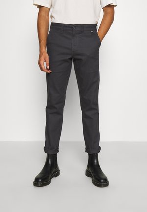 Mann trägt dunkelgraue Slim-Fit-Hosen mit aufgekrempelten Bündchen und schwarze Leder-Stiefeletten, steht mit entspannt an den Seiten hängenden Händen.