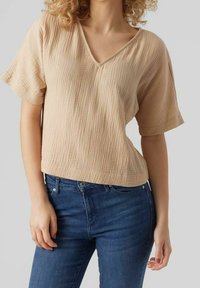 Haut beige à manches courtes avec un col en V, présentant un motif texturé. Associé à un jean bleu foncé, mettant en valeur son design décontracté.