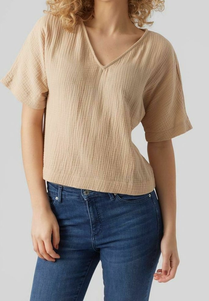 Haut beige à manches courtes avec un col en V, présentant un motif texturé. Associé à un jean bleu foncé, mettant en valeur son design décontracté.